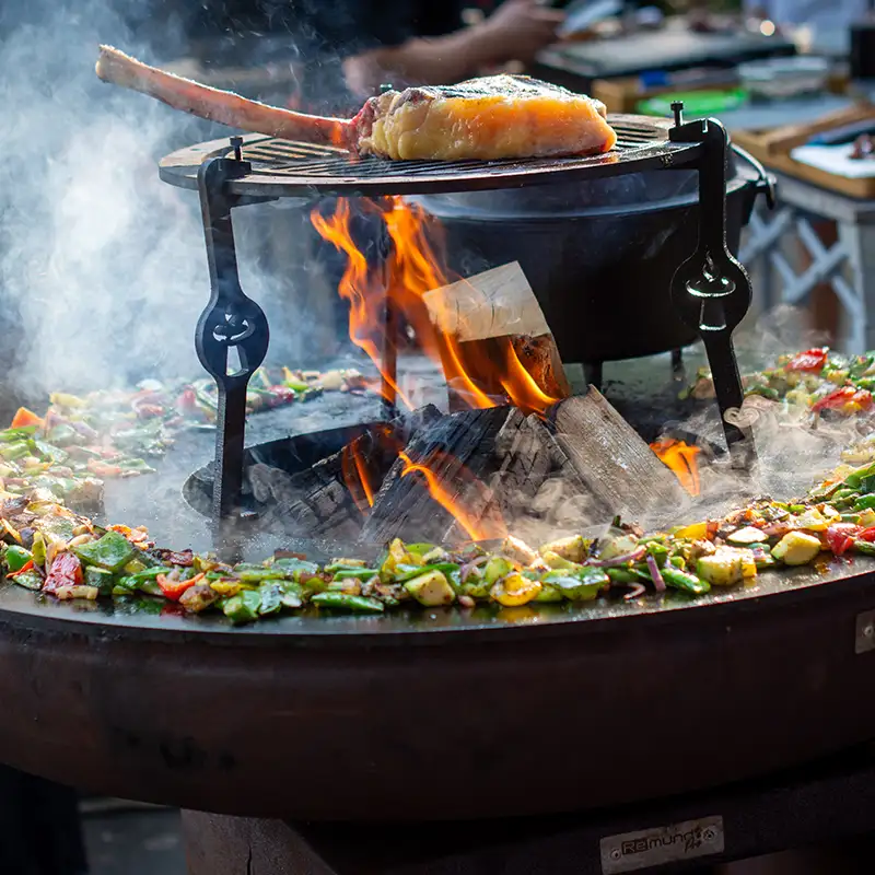 Remundi Carus Feuerstelle voll belegt mit verschiedenem Grillgemüse und dem Topper-Aufsatz, auf dem ein Stück Fleisch über offener Flammen liegt