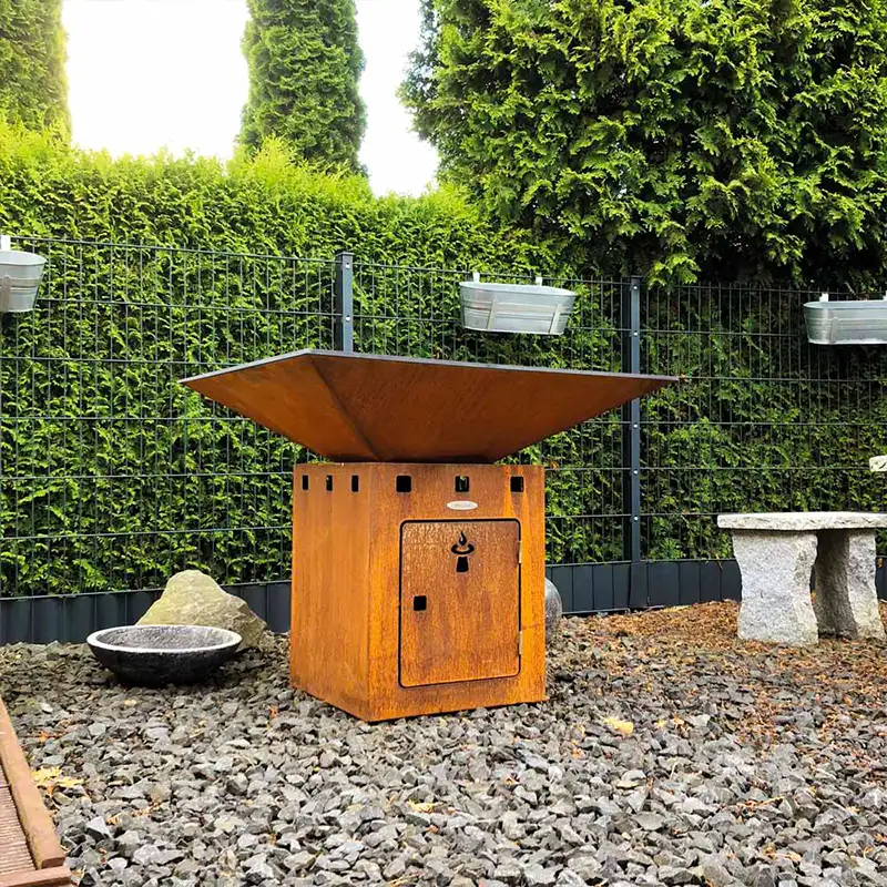 Eckige Feuerstelle steht auf einem mit kleinen Steinen belegten Boden in einem Garten