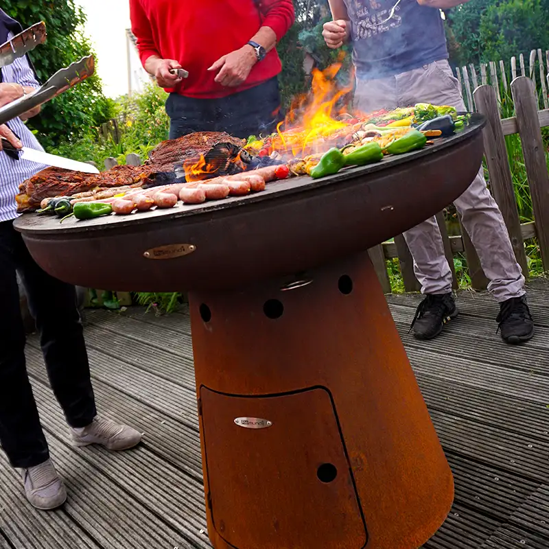 Mehrere Personen stehen vor der Remundi Nero Feuerstelle in einem Garten und braten Grillgut
