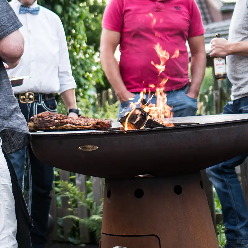 Mehrere Leute stehen vor einer befeuerten Remundi Nero Feuerstelle, auf der Fleisch gegrillt wird
