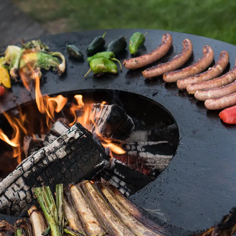 Detailansicht von der runden Plancha-Platte mit Feuerstelle und gebratenen Würstchen und Gemüse