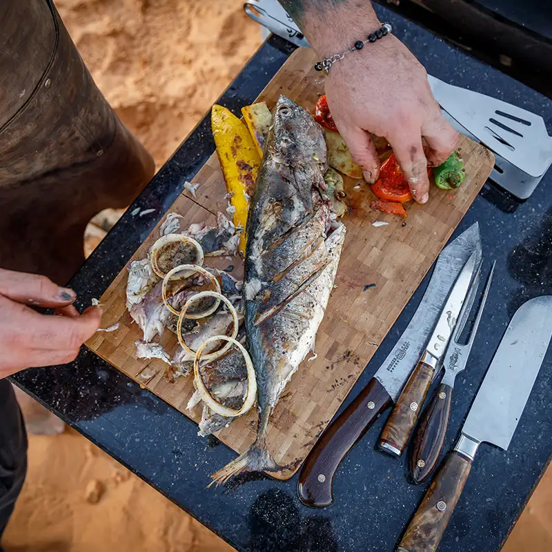 Schneidebrett auf dem ein gebratener Fisch und Gemüse liegen, daneben mehrere Grillutensilien