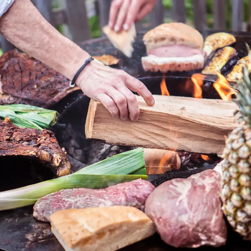 Eine Person legt ein großes Stück Feuerholz in die Mitte der Remundi Feuerstelle, auf der Grillplatte liegt verschiedenes rohes Grillgut