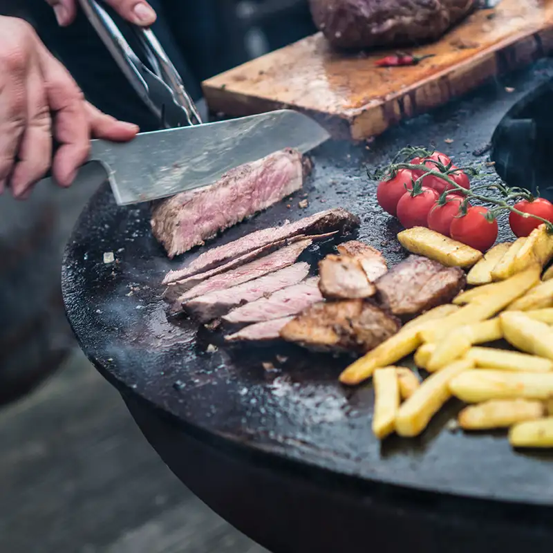 Jemand schneidet mit einem Messer ein fertig gegrilltes Steak in feine Streifen direkt auf der Remundi Grillplatte