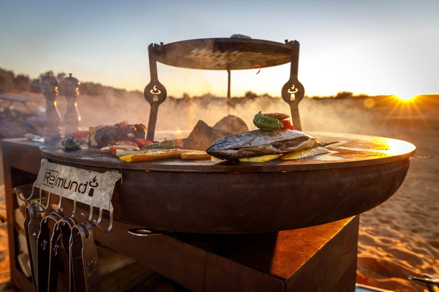 Feuerstelle Vulcano L von der Seite atmosphärisch beleuchtet. Auf dem Grill liegen Fisch und Gemüse.