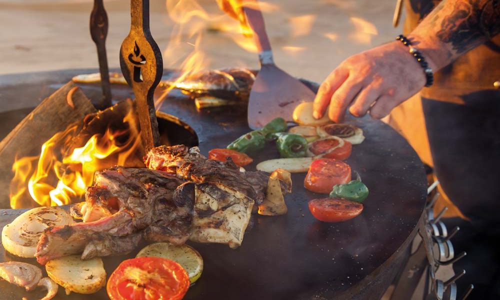 Remundi Feuerplatte im Einsatz mit verschiedenem Grillgut: Gemüse und Fisch auf dem Stahlring, während in der Mitte modulares Zubehör genutzt wird.