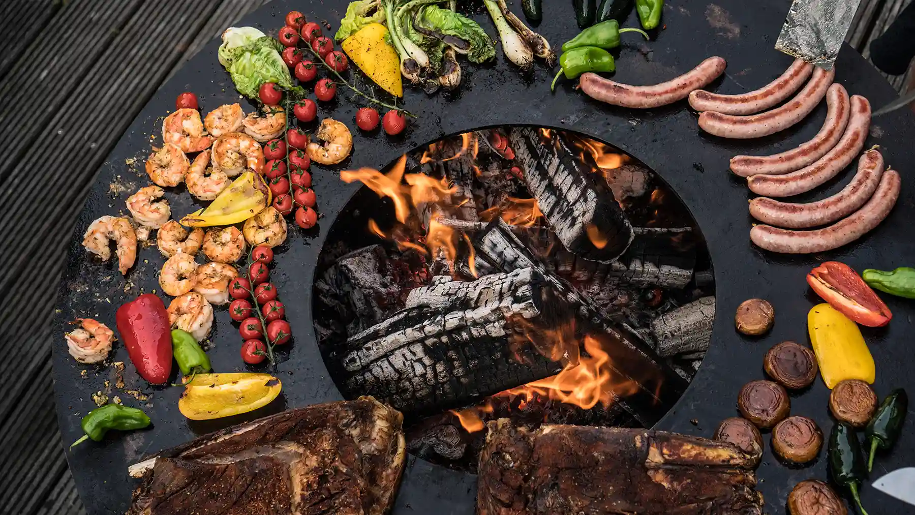 Ansicht von oben auf runde Feuerplatte mit Feuerholz, gegrilltem Fleisch, Fisch und Gemüse